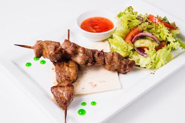 Shish kebab on bamboo skewers lies on a pita. Near sauce and fresh vegetable salad. White background (close)