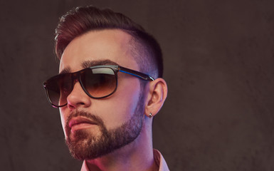 Close-up portrait of a confident stylish bearded man with hairstyle and sunglasses in a gray suit and pink shirt, posing in a studio.