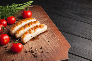 Grilled sausages grilled on a brown wooden board on a wooden background, six cherry tomatoes, greens, parsley dill, pepper, sauce. Place under the text and logo. Top view. Flat lay.