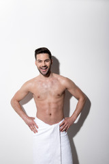 Happy smile. Portrait of cheerful young bearded man is standing in towel and looking at camera with joy. He is holding hands akimbo while demonstrating his muscular body
