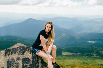 Naklejka na ściany i meble Young girl sitting on stone in mountains. Mood portrait of long haired adorable blonde babe outdoor on vacation. Female hiker relaxing at nature. Beautiful landscape view. Cute lovely model posing.