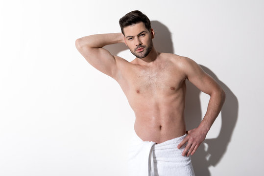Portrait of pensive young bearded man is standing with towel on his hips and looking at camera thoughtfully. He is holding hand behind his head and other one on his hip. Copy space in the left side