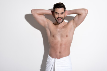 Fototapeta premium Enjoying my life. Portrait of masculine young smiling man with stubble is standing in towel and looking at camera with joy while holding hands behind head. Beauty concept