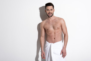 Positiveness. Portrait of cheerful young bearded man in towel is standing against white wall and looking at camera with joy. Copy space in the left side. Beauty concept
