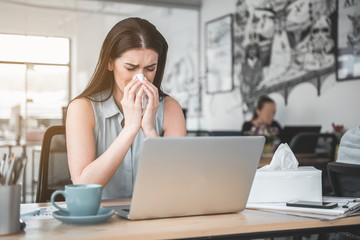 Portrait of frustrated girl blowing nose into handkerchief while working with laptop. Unhappy sick worker concept