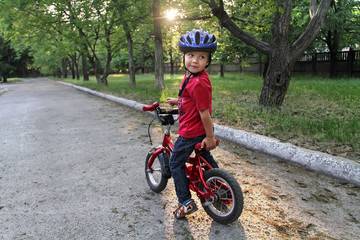 A small white Toddler boy in a protective helmet on his head sits on a children's bicycle. Toddler on a two-wheeled red bicycle looks back. A sly smile on the child's face