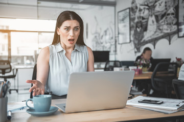 What is it. Amazed female watching at notebook computer while sitting at table in office. Worried employer concept