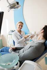Obraz premium Portrait of smiling female dentist holding tooth model and showing how to brush teeth to unrecognizable patient sitting in dental chair