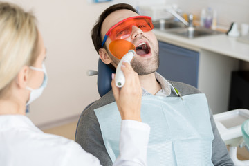 Portrait of mature man sitting in dental chair with mouth open with female dentist performing laser...