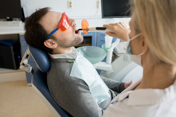 Side view portrait of mature man sitting in dental chair with mouth open while female dentist performing laser teeth whitening in clinic, copy space