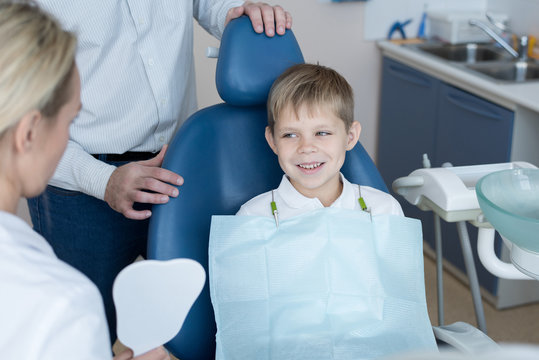 Portrait of smiling  little boy sitting in dental chair  listening to female dentist holding tooth model explaining oral hygiene rules with father at his side