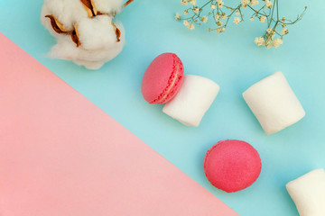 Top view of mini pink macaron or macaroon french desserts cake with marshmallows and cotton flowers on soft sweet pink and blue pastel geometric paper flat lay background.