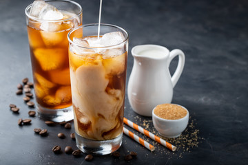 Ice coffee in a tall glass with cream poured over, brown sugar and coffee beans. Cold summer drink on a dark background. With copy space