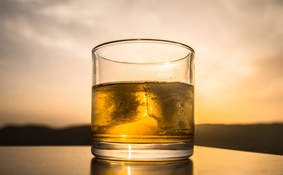 A glass of whiskey with ice on a sunset background or shot of whiskey at sunset dramatic sky on mountain landscape background