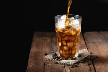 Ice coffee in a tall glass over and coffee beans on a old rustic wooden table. Cold summer drink on a dark background with copy space. The process of pouring drink from a coffee pot into a glass