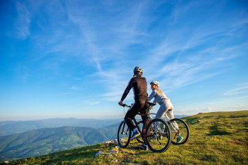 Naklejka na ściany i meble Cyclist couple with mountain bikes standing on the hill under the evening sky and enjoying bright sun at the sunset.