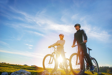 Cyclist couple with mountain bikes standing on the hill under the evening sky and enjoying bright sun at the sunset.