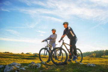 Fototapeta na wymiar Cyclist couple with mountain bikes standing on the hill under the evening sky and enjoying bright sun at the sunset.