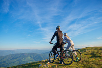 Naklejka na ściany i meble Cyclist couple with mountain bikes standing on the hill under the evening sky and enjoying bright sun at the sunset.