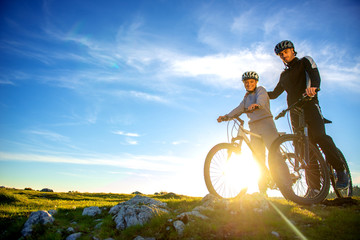 Fototapeta na wymiar Cyclist couple with mountain bikes standing on the hill under the evening sky and enjoying bright sun at the sunset.