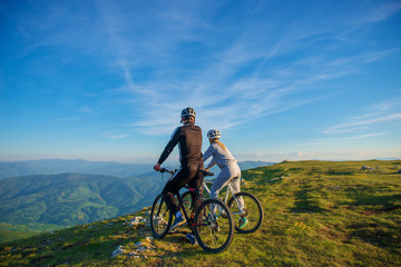 Fototapeta premium Cyclist couple with mountain bikes standing on the hill under the evening sky and enjoying bright sun at the sunset.