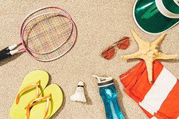 top view of badminton equipment, sunglasses, flip flops, cap and towel on sand
