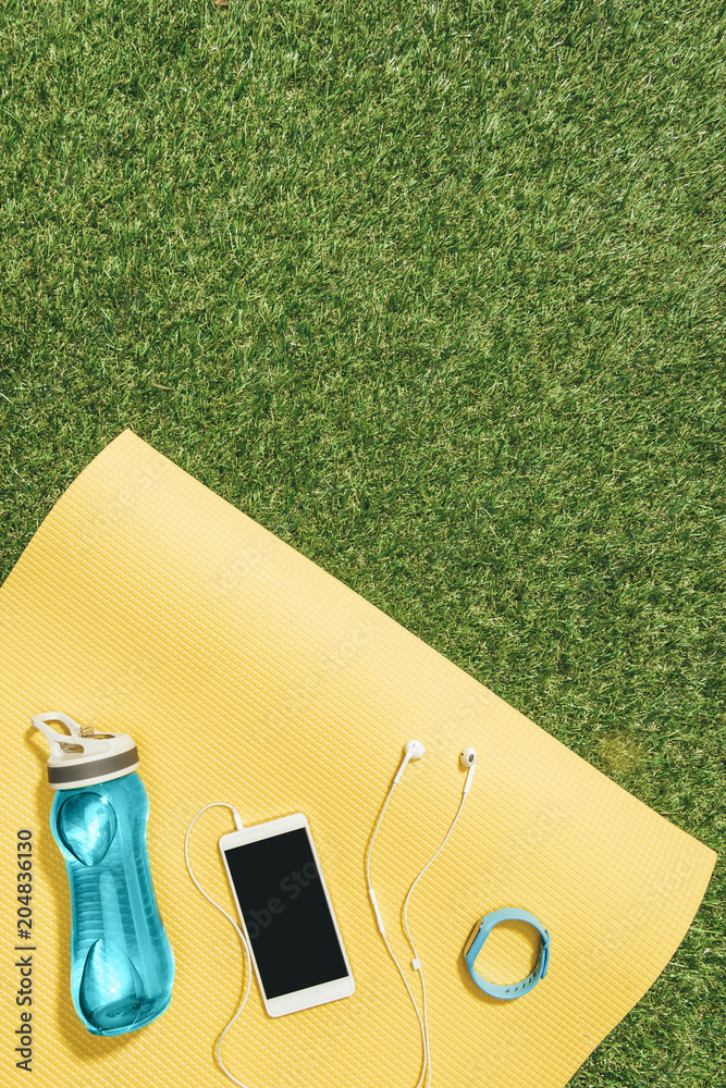 Wall mural flat lay with water bottle, watch, smartphone and earphones on yellow mat on green lawn