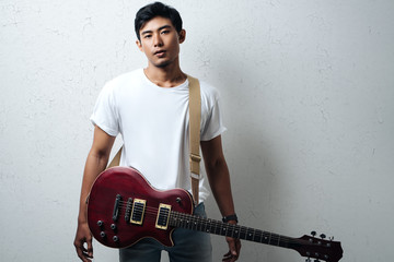 Handsome guy with guitar and white blank t-shirt, grunge wall, horizontal studio portrait