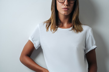 Pretty woman in white blank t-shirt wearing glasses, empty wall, horizontal studio close-up