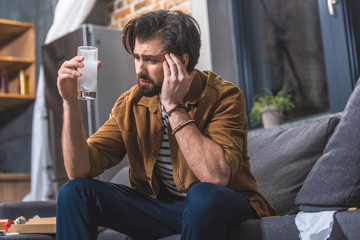 loner having headache and hangover and looking at glass of water with pill at living room