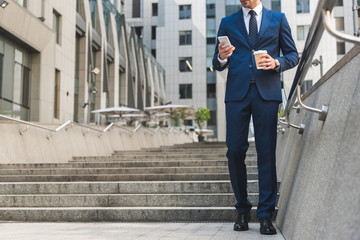 cropped shot of businessman in stylish suit with coffee to go using smartphone while going down...