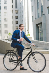 handsome young businessman with paper cup of coffee on bicycle in business district