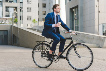 young businessman in stylish suit with coffee to go sitting on vintage bicycle