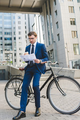 Fototapeta na wymiar businessman in stylish suit with newspaper and paper cup of coffee leaning on bicycle