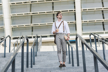attractive young man walking down stairs in front of stadium with book and coffee to go