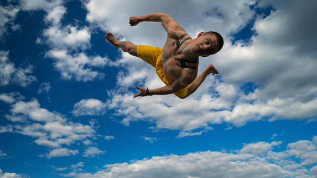 Tricking on street. Martial arts. Man performs somersault ahead barefoot. Shooted from bottom foreshortening against sky.