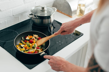 cropped shot of man cooking vegetables in frying pan on electric stove - obrazy, fototapety, plakaty