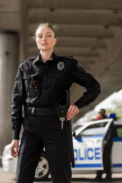 attractive female police officer in uniform looking at camera with blurred partner near car on background
