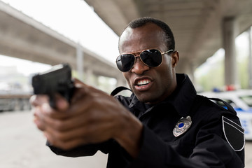 angry african american policeman shouting and aiming by handgun