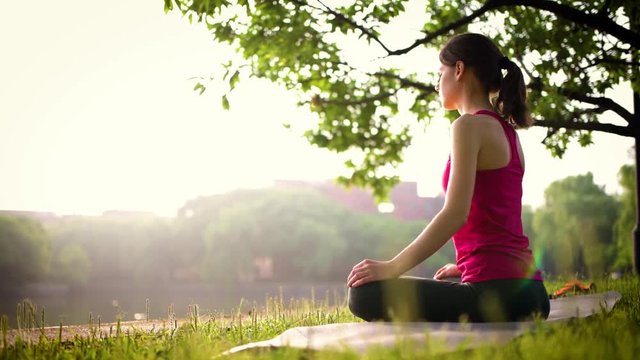 Beautiful girl doing yoga and practicing meditation in the beautiful park near the lake in the sunlight, sunrise