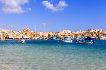 Fototapeta na wymiar Marsaxlokk historic port with many boats in transparent sea, Malta. Blue sky and village background.