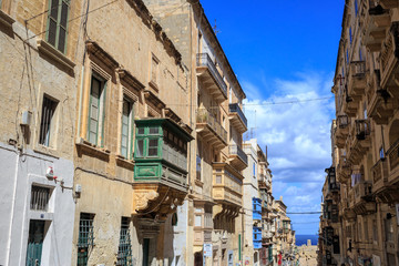 Fototapeta na wymiar Malta, Valletta, traditional sandstone buildings with colorful wooden windows on balconies. Blue sky with clouds and sea background.