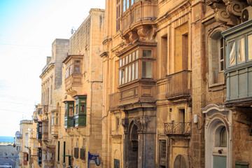 Malta, Valletta, traditional sandstone buildings with colorful wooden windows on balconies. Blue sky with clouds and sea background.