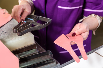 Сlose-up of a young woman working as a seamstress in a purple unifrome cuts out pink genuine leather children's shoes on a table.