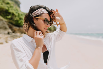 Adorable lovely woman with dark wavy hair dressed white shirt and black sunglasses has fun on the white beach near the ocean with lovely smile. Recreation, vacation mood, summer, sunny day