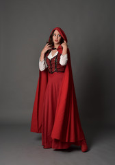 Naklejka premium full length portrait of woman wearing red fantasy costume with cloak, standing pose on grey studio background.