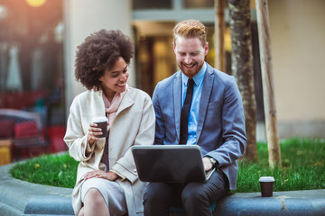 Two business people in an informal conversation in front of a business building using laptop and...