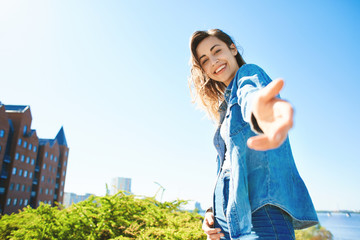 portrait of a young smiling attractive woman in jeans clothes at sunny day on the blue sky background. happy woman gives a hand to someone like follow me . first person view. - obrazy, fototapety, plakaty