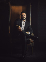 Przystojny mężczyzna z brodą w klasycznym garniturze siedzi na skórzanym krześle i patrzy w kamerę. - 204864948