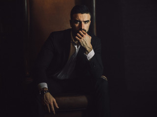 A handsome man with a beard and in a classic suit sits in a leather chair and looks at the camera. 